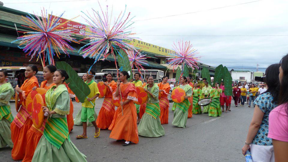 Eine Gruppe von Menschen in bunten Kostümen marschiert auf einer Straße während eines Karnevalsumzugs, wobei eine Frau rechts eine Flasche hält, und Fahrzeuge, Gebäude, Bäume, Pfosten, Drähte und der Himmel im Hintergrund zu sehen sind.