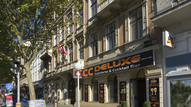 Eine belebte Stadtstraße mit verschiedenen Gebäuden, Fahrzeugen, Fußgängern und Schildern, mit einem hohen Gebäude mit der Aufschrift "CC Deluxe Cafe & Cocktail Bar" in der Mitte.