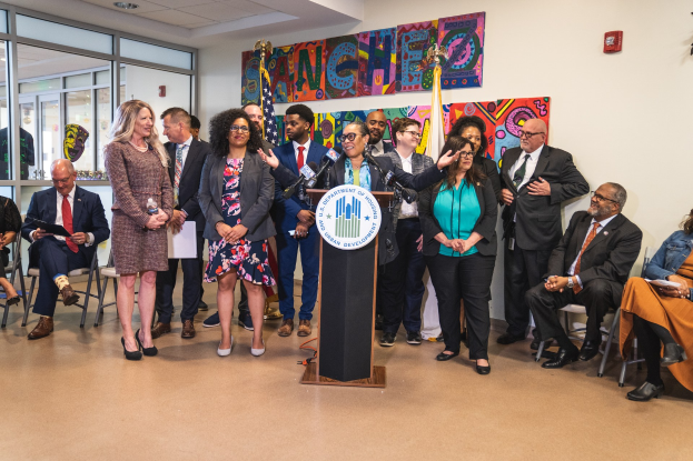 Gruppe von Menschen vor einem Rednerpult mit Mikrofon und einer Tafel mit Text, einige sitzen auf Stühlen und andere stehen, mit Flaggen, einer Tafel, einem Feuerlöscher und Deckenlampen an der Rückwand.