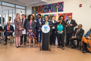 Gruppe von Menschen vor einem Rednerpult mit einem Mikrofon und einer Tafel mit der Aufschrift "Sancho-Stadtrat tritt dem Stadtrat bei", mit sitzenden und stehenden Teilnehmern, Flaggen, einem Feuerlöscher und Deckenleuchten im Hintergrund.