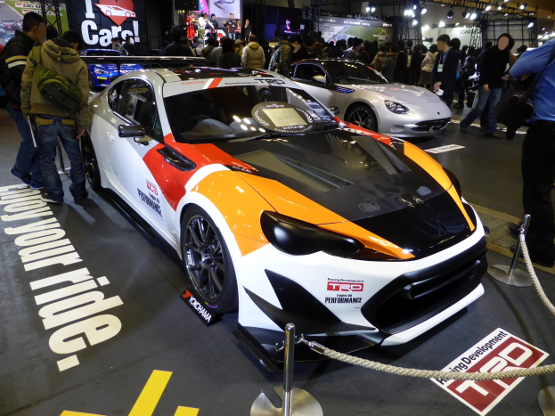 Eine Gruppe von Menschen betrachtet ein Rennfahrzeug auf der Tokyo Motor Show, umgeben von Absperrpollern mit Seilen, mit Informationsschildern im Hintergrund unter beleuchteter Decke.