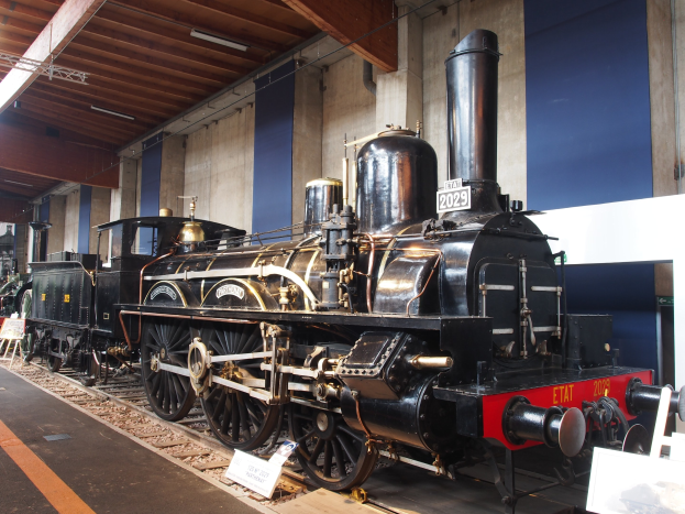 Alte Dampflokomotive in einem Museums-Setting mit umgebenen Eisenbahnschienen, Informations-Tafeln und anderen Ausstellungsstücken an einer Wand mit Deckenbeleuchtung.