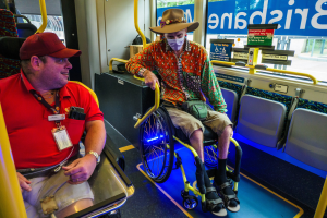Zwei Männer im Rollstuhl in einem Bus, einer trägt ein rotes T-Shirt und eine Kappe, der andere ein orangefarbenes Hemd und einen braunen Hut, beide tragen Masken und Ausweise, mit gelben Stangen und Glasfenstern, durch die man Bäume, Gebäude und den Himmel sieht.