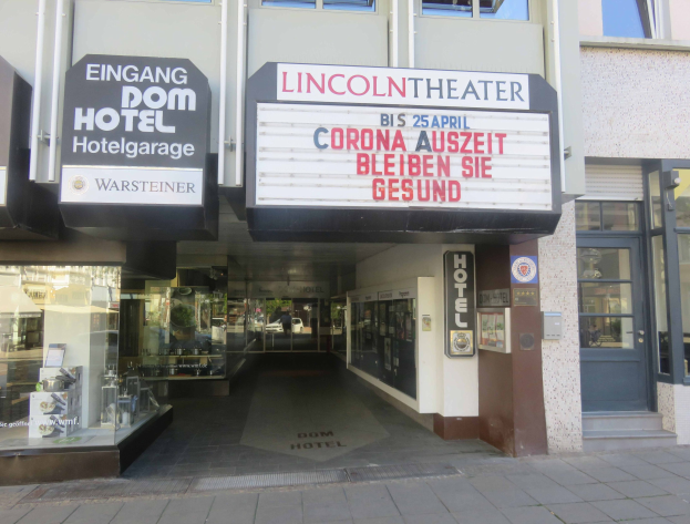 Außenansicht des Lincoln Theaters in Berlin, Deutschland, mit Glasfenstern und -türen sowie einer Tafel und einem Innenraum, der eine belebte Stadtlandschaft suggeriert.