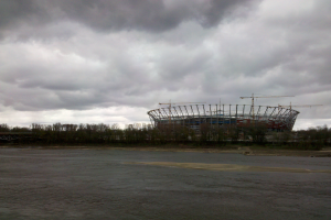 Das Olympiastadion in Kiew, Ukraine, ein großer Stadionkomplex umgeben von Bäumen und einer Brücke, mit bewölktem Himmel im Hintergrund und sichtbarem Boden unten.