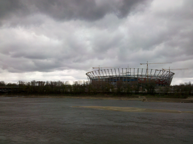 Das Olympiastadion in Kiew, Ukraine, ein großer Stadionkomplex umgeben von Bäumen und einer Brücke, mit bewölktem Himmel im Hintergrund und sichtbarem Boden unten.