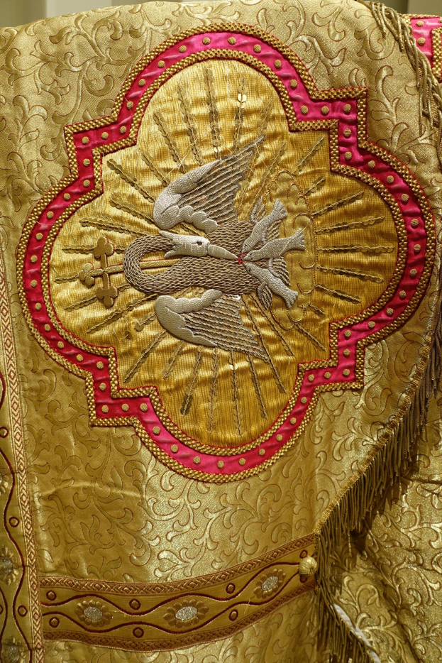 Nahaufnahme eines Priestergewandes mit aufwendiger Gold- und Roterstickerei vor einer Wand im Hintergrund.