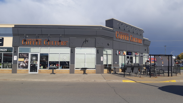 Ein Kaffeeshop an einer Straßenecke mit einer Straße im Vordergrund, einem Gebäude im Hintergrund, einigen Fahrzeugen, Bäumen und einem blauen und weißen Himmel.