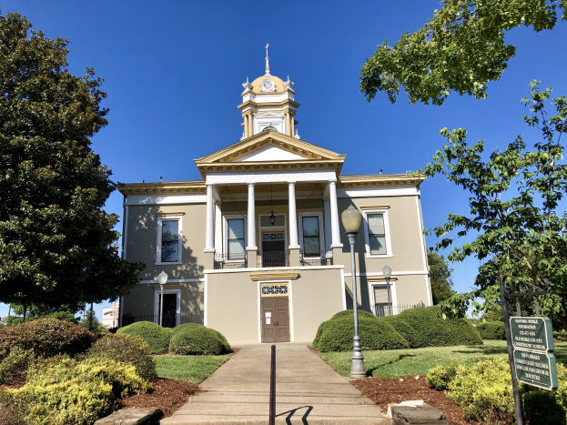 Großes Backsteingebäude mit einem Uhrenturm, mehreren Fenstern, Türen und Säulen, umgeben von Straßenmöbeln, Grünflächen und einem bewölkten Himmel, als das wiedereröffnete alte Rathaus bezeichnet.