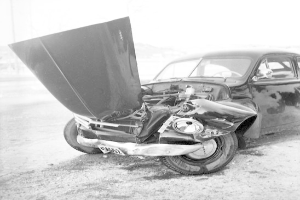 Ein stark beschä-digtes Schwarz-Weiß-Foto eines Wagens, der am Straßenrand liegt, mit eingedellter Motorhaube, zerbrochenen Fenstern und einer offenen Fahrertür, die den Motor freilegt.