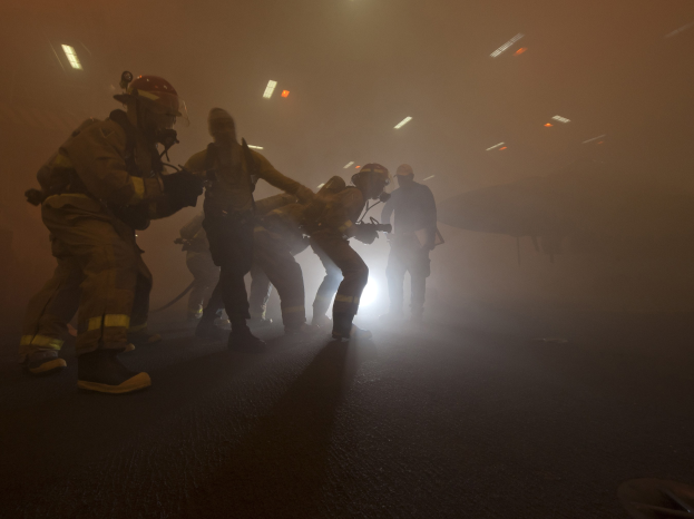 Feuerwehrleute in Helmen gehen auf einer nebligen Straße und tragen Gegenstände, mit einem Flugzeug und Lichtern im Hintergrund und einer Straße unten.