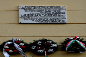 Zwei Kränze mit Bändern hängen an der Seite eines Gebäudes, mit einer Gedenktafel für Holocaust-Opfer an der Wand dahinter.