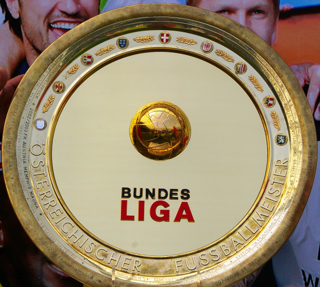 Ein Mann hält einen goldenen Schild mit der Aufschrift 'Bundesliga', im Hintergrund eine Fahne mit drei Personen