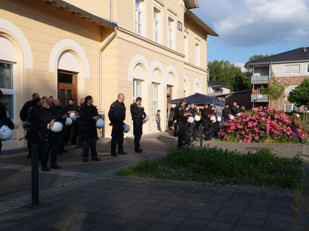 Eine Gruppe von Polizisten vor einem Gebäude mit Fenstern und Türen, die Helme halten, in der Nähe eines Zeltes, umgeben von Pflanzen, Blumen, Gras, einem Straßenpfahl, Bäumen und einem bewölkten Himmel.