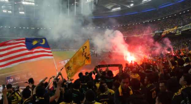 Eine Menschenmenge in einem Stadion mit Fackeln, Fahnen und einer Kamera, mit Rauch und Schildern, die "Malaysia vs. Malaysia Live-Streaming" im Hintergrund zeigen.