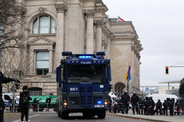 Gruppe von Polizeibeamten vor einem großen Gebäude mit Fenstern, Säulen und Bögen, mit Fahrzeugen auf der Straße und einer Person mit einer Kamera auf der linken Seite, Bäume und Verkehrszeichen im Hintergrund unter einem klaren blauen Himmel.