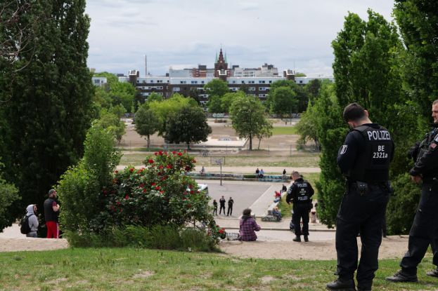 Zwei Polizeibeamte vor einer Gruppe von Menschen in einem Park mit saftigem Grün, Bäumen, bunten Blumen, Gebäuden, Pfählen und einem klaren blauen Himmel.