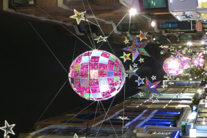 Eine Straßenlaterne in einer nächtlichen Stadt mit zahlreichen Lichtern und Dekorationen, darunter eine große Diskokugel, die von einem Gebäude hängt und eine festliche Atmosphäre schafft.