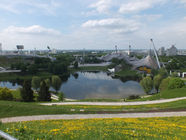 Blick auf das Olympiastadion von einem Hügel aus, mit einem Teich umgeben von grünem Gras, Bäumen und gelben Blumen im Vordergrund und Gebäuden und einem klaren Himmel im Hintergrund.