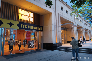 Kino mit Glaswänden und Eingangstreppe, Menschen vor dem Gebäude, beleuchtet von Lichtern, mit Informationsschildern an den Wänden, Bäumen und blauem Himmel im Hintergrund und einem Wasserzeichen in der rechten unteren Ecke.