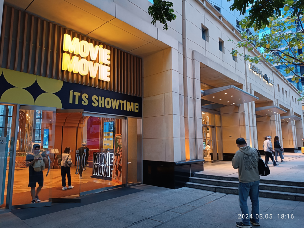 Kino mit Glaswänden und Eingangstreppe, Menschen vor dem Gebäude, beleuchtet von Lichtern, mit Informationsschildern an den Wänden, Bäumen und blauem Himmel im Hintergrund und einem Wasserzeichen in der rechten unteren Ecke.