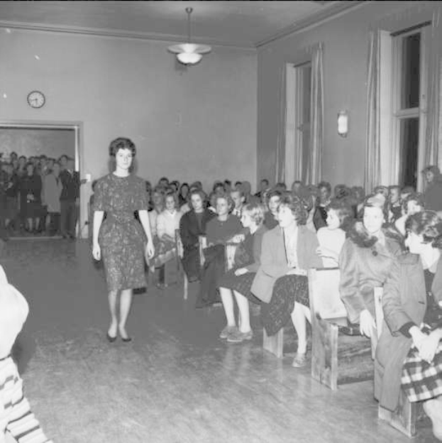 Eine Frau in einem schwarzen und weißen Kleid läuft über einen Laufsteg während einer Modenschau für Frauenwahlrecht, mit einer Menge, die auf Stühlen sitzt und einer Uhr und Lichtern an der umliegenden Wand.
