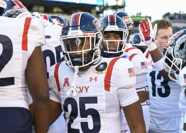 Gruppe von Männern in Football-Outfits auf einem Feld mit Helmen und Handschuhen, mit einer unscharfen Menge, einer Anzeigetafel mit 'Auburn vs Virginia Tech - 11/28/15 College Football Pick, Odds, and Prediction' und Himmel im Hintergrund.