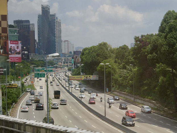 Eine vielbefahrene Autobahn mit hohen Gebäuden, Straßenlaternen, Werbetafeln, Bäumen und bewölktem Himmel.