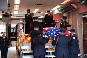 Eine Gruppe uniformierter Männer trägt einen Sarg mit einer amerikanischen Flagge darauf, umgeben von ein paar Männern mit Mützen und einem Löschfahrzeug und Deckenlichtern im Hintergrund.