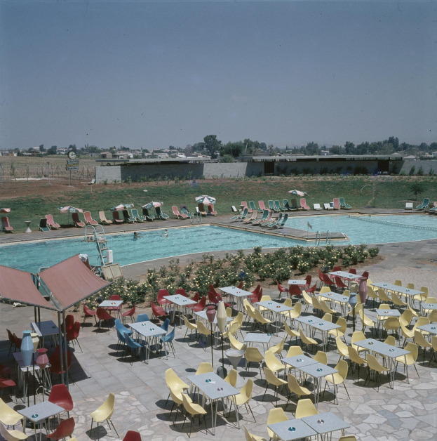 Ein Freibad mit Tischen, Stühlen und Sonnenschirmen umgeben von Pflanzen, einem Zaun, einer Wand, Bäumen, Gebäuden und einem bewölkten Himmel.