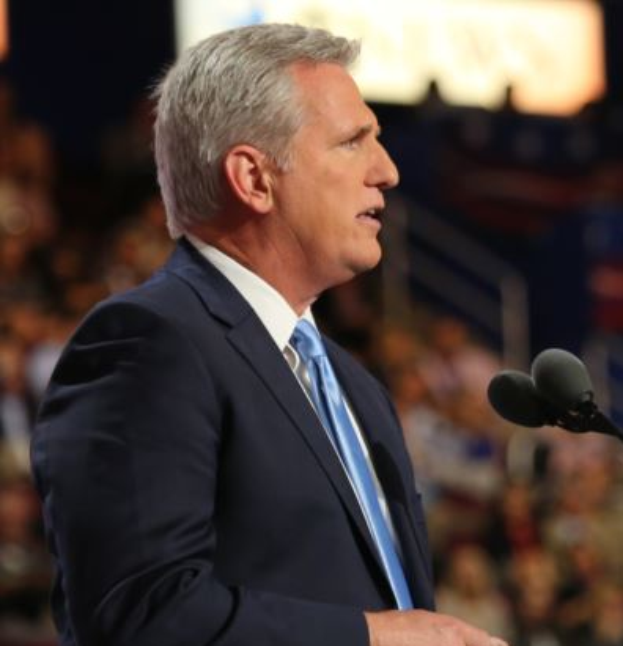 Ein Mann im Anzug und mit Krawatte steht an einem Podium mit zwei Mikrofonen und sieht ernst aus, mit einem unscharfen Hintergrund.