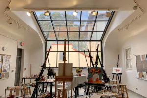 Ein unordentliches Kunstatelier mit zahlreichen Gemälden auf Staffeleien, Hockern, Tischen, Vasen, Brettern, Papieren, Büchern und anderen Gegenständen, das eine Glaswand mit Blick auf Bäume, Gebäude und Himmel, ein Anschlagbrett mit Papieren und eine Uhr auf der linken Seite und Fenster auf der rechten Seite hat und Teil des neu eröffneten Studios des Art Institute of Chicago ist.