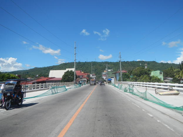 Autobahn mit fahrenden Fahrzeugen, ein geparktes Motorrad auf dem Seitenstreifen, Leitplanken, Strommasten mit Drähten, Bäume, entfernte Gebäude, Hügel und ein bewölkter Himmel.