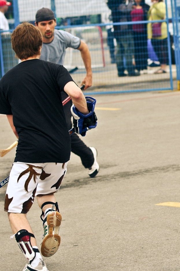 Zwei Menschen, die Hockey auf einer Straße mit einem Zaun dahinter und einer Menge von Menschen im Hintergrund spielen.