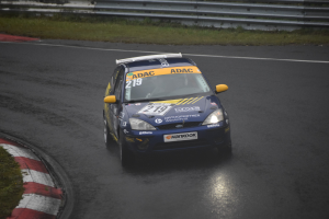 Ein blaues Rennauto mit Text und Zahlen darauf fährt auf einer Strecke, flankiert von Gras und Geländern, mit Bäumen im Hintergrund.