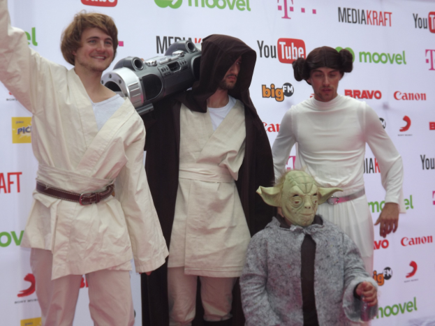 Eine Gruppe von Menschen in Star-Wars-Kostümen, die auf einem roten Teppich posieren und im Hintergrund ein Banner mit Text und Logos zu sehen ist.