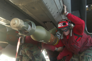 Eine Gruppe von Männern in roten Jacken und Tarnuniformen arbeitet an einem Flugzeug, wobei einer einen Helm, eine Schutzbrille und eine Uhr trägt und im Hintergrund eine Rakete zu sehen ist.