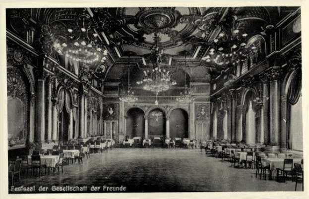 Schwarz-weiß-Foto eines großen Raums mit Tischen, Stühlen, Säulen, Bögen und Kronleuchtern, beschriftet mit "Festival der Gesellschaft der Freunde, Berlin" unten.