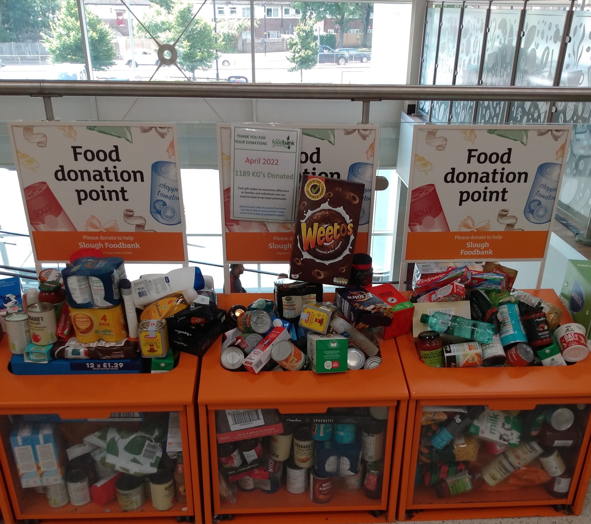 Orange Schübe mit Lebensmittelspenden gefülllt, darunter Flaschen, Dosen und Kisten, mit Informationsschildern; vor dem Glasfenster sind Fahrzeuge, Bäume, Gebäude und Himmel zu sehen.