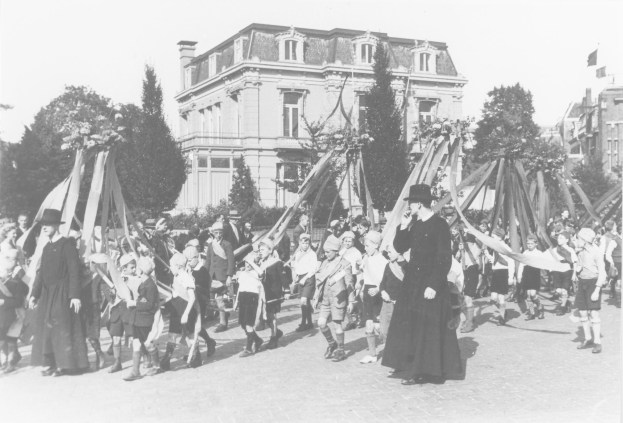 Eine Gruppe von Menschen in Schwarz-Weiß-Kleidung marschiert mit Fahnen durch eine Straße, mit Bäumen und Gebäuden im Hintergrund und einem Maibaum.