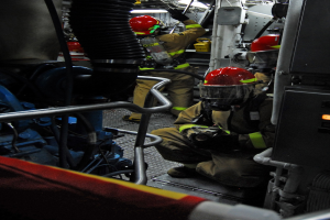 Feuerwehrleute in Schutzausrüstung arbeiten an einem Löschfahrzeug mit Rohren und einer Lampe im Hintergrund.