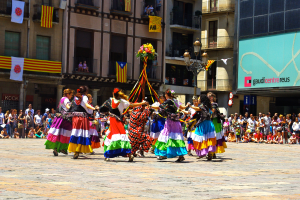 Eine Gruppe von Frauen in bunten Kleidern tanzt auf der Straße, umgeben von einer Menge Menschen, mit Gebäuden, Gelöndern, Balkonen, Fahnen, einem Lichtmast und einer Tafel mit Aufschrift im Hintergrund.
