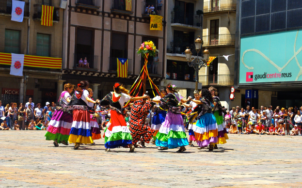 Eine Gruppe von Frauen in bunten Kleidern tanzt auf der Straße, umgeben von einer Menge Menschen, mit Gebäuden, Gelöndern, Balkonen, Fahnen, einem Lichtmast und einer Tafel mit Aufschrift im Hintergrund.