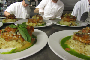 Drei Köche in einer Küche mit Mützen und Handschuhen bereiten Essen auf Tellern mit verschiedenen Gegenständen vor, mit Regalen voller Vorräte im Hintergrund.