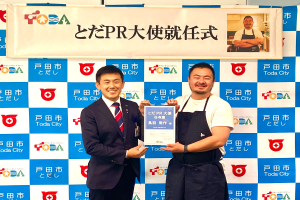 Zwei strahlende Männer mit einem Zertifikat, im Hintergrund ein Banner mit der Aufschrift "Toda City Wins PR Award" und einem Foto einer Person.