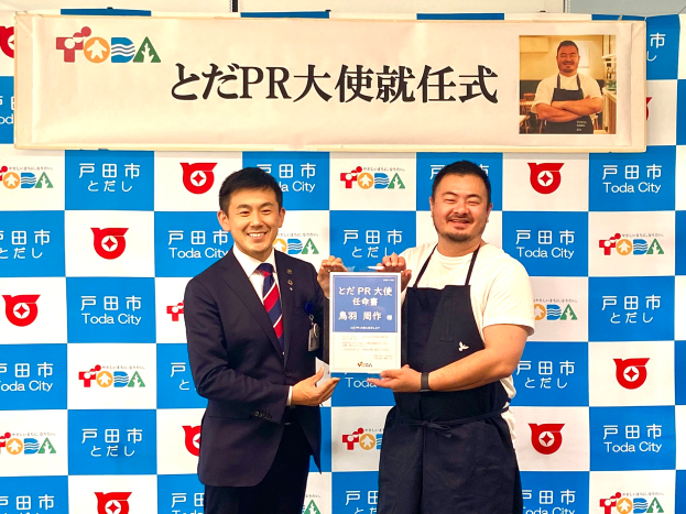 Zwei strahlende Männer mit einem Zertifikat, im Hintergrund ein Banner mit der Aufschrift "Toda City Wins PR Award" und einem Foto einer Person.