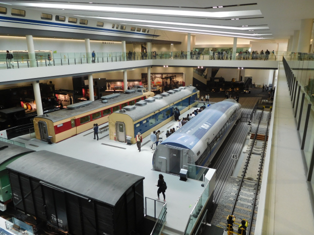 Ein großer Museumsraum mit verschiedenen Bahnen in unterschiedlichen Größen und Farben, mit Menschen, die beobachten, Geländern, Säulen und Deckenleuchten.