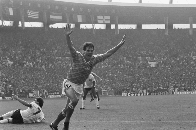 Schwarzes und weißes Foto eines Fußballspielers, der nach einem Tor jubelt, umgeben von Teamkollegen und Stab auf dem Feld, mit Stadiontafeln, Fahnen und Pfosten im Hintergrund.