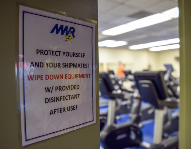 Ein Türschild mit der Aufschrift "Schützen Sie sich und Ihre Schiffskameraden" mit Laufbändern und Deckenleuchten im Hintergrund, mit einem leicht unscharfen, bewegungartigen Effekt.