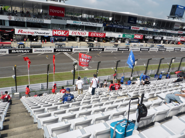 Eine Gruppe von Menschen sitzt auf der Tribüne einer Rennstrecke, mit Fahnen, Schildern, Geländern und Stufen, vor einem bewölkten Himmel.